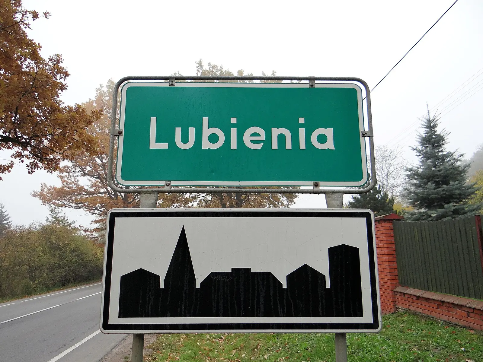 Tablica z nazwą miejscowości Lubienia przy drodze, w tle jesienne drzewa i ceglany mur
